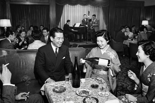 白黒。男性にお酌する和装の女性。