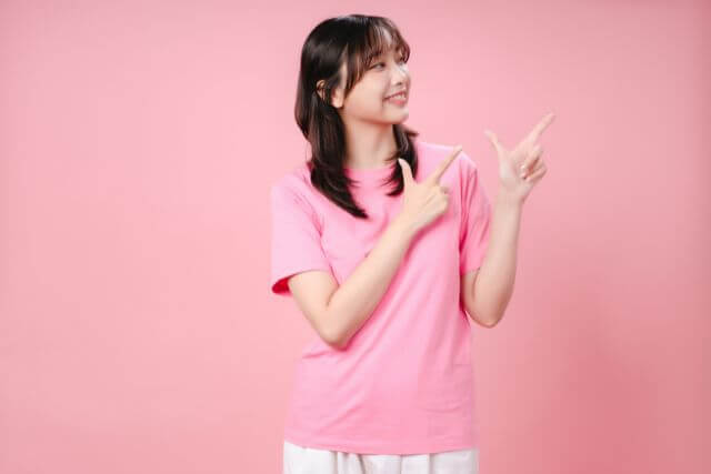 ピンクの壁の前でピンクのT札を着て両手の人差し指を立てている若い女性