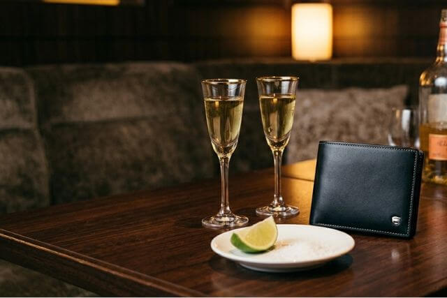 テキーラが注がれた2杯のグラスと、男性用の高級なノーブランドの財布がテーブルに置かれた様子