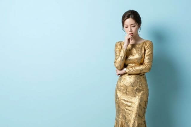 水色の壁の前に立って顎に手を当てて俯くゴールドのドレスを着た若い女性