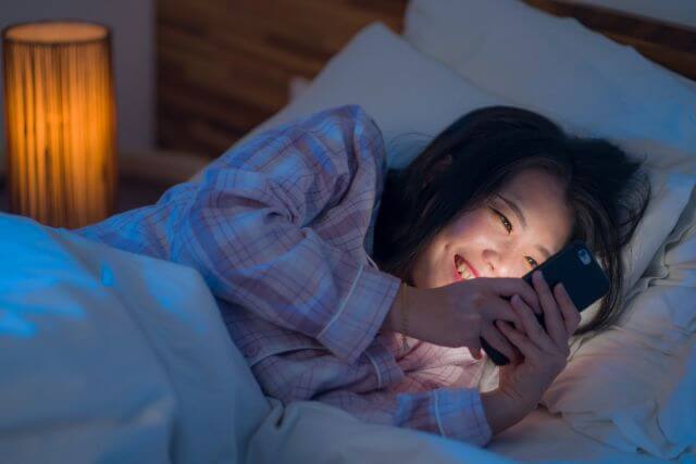 夜にベッドの中でスマホを見て笑顔の女性