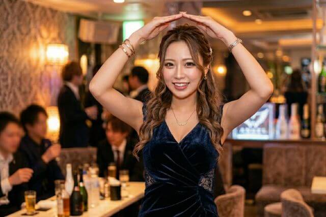 キャバクラ店内で頭上で丸を作るロングヘアのキャバ嬢
