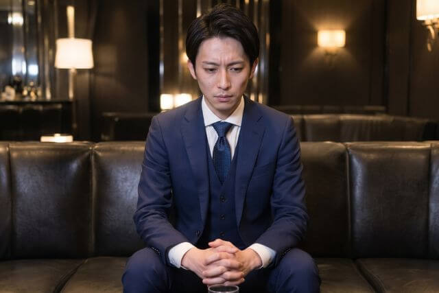 スーツの男性が1人でソファに座っている。伏し目がちで緊張しているような感じ