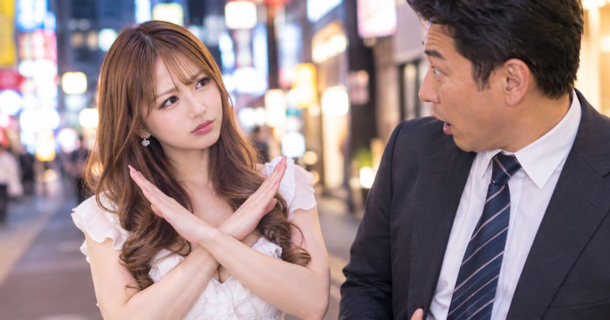 白いフリルのトップスを着た女性が、胸の前で腕をクロスしてバツ印を作って困った表情をして男性の方を見ている。隣のスーツの男性が驚いた表情で女性を見ている。