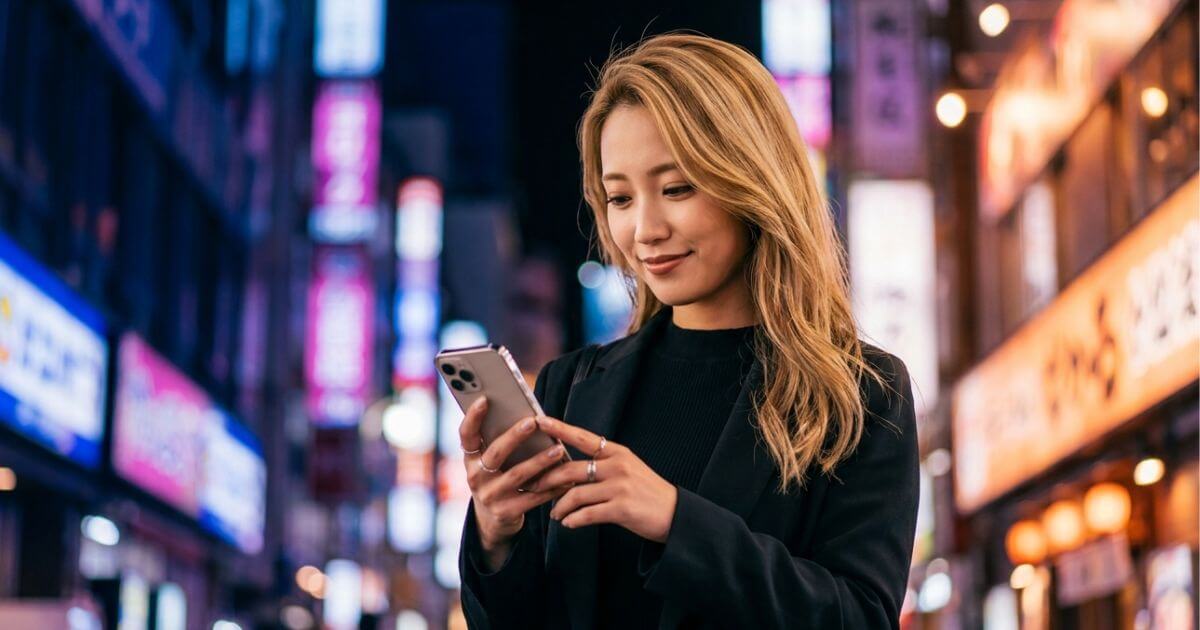 夜の繁華街でスマホを見ているブロンドヘアの若い女性